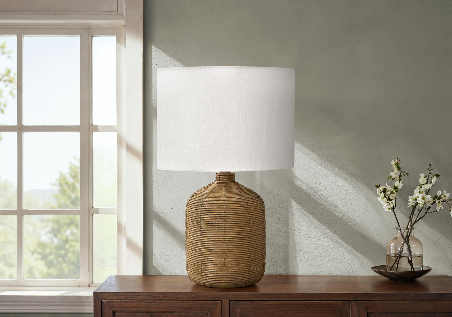 A modern rattan table lamp sits on a wooden console by the window, accompanied by a small vase of white flowers and a decorative bowl against a light gray wall.