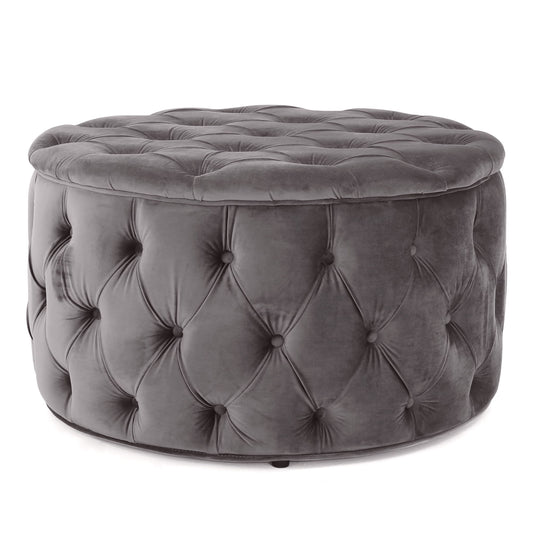 The Modern Glam Ottoman Velvet is a round ottoman, upholstered in dark gray velvet with a plush cushioned top and button-tufted sides.