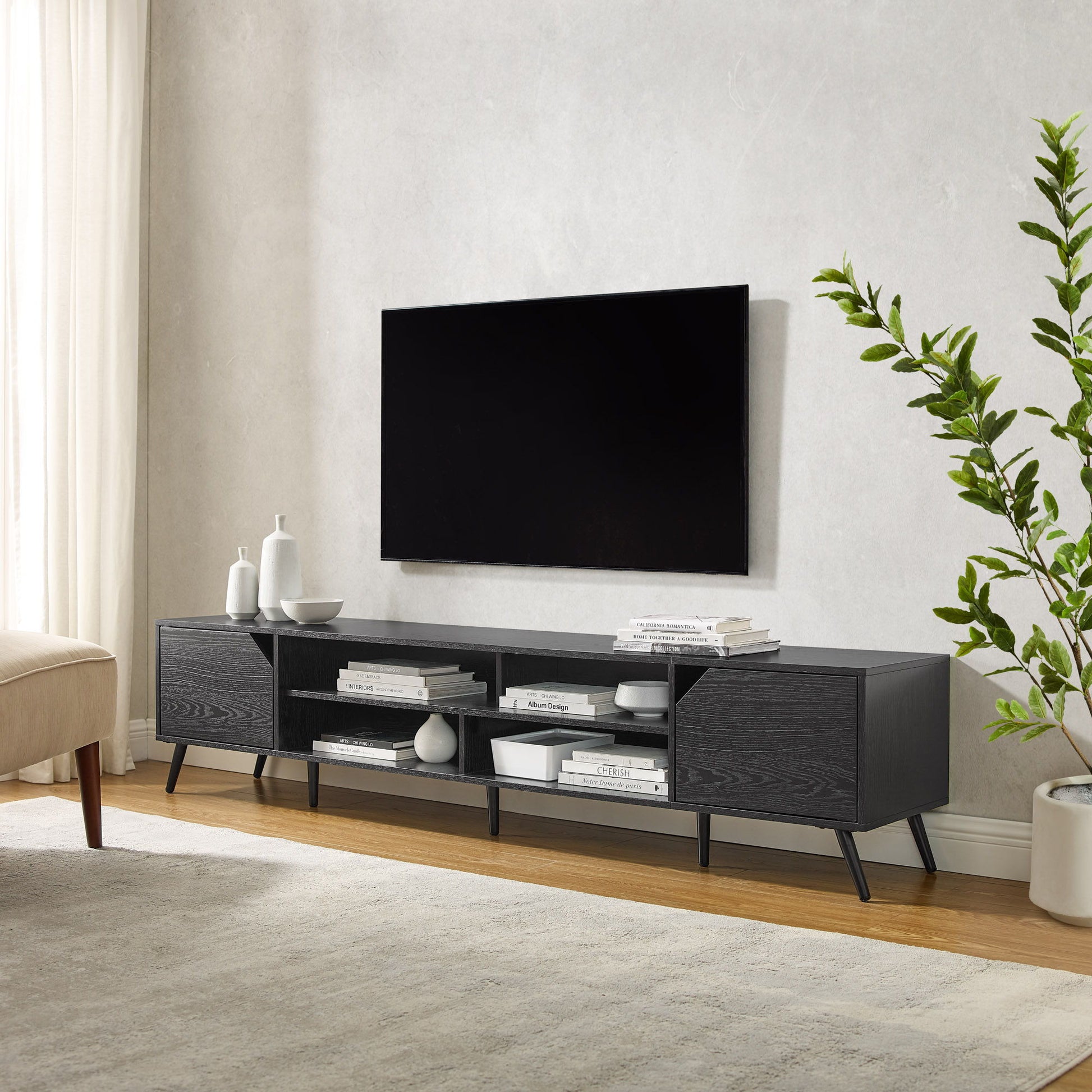 A modern living room with a wall-mounted TV above the Contemporary 2 Door Minimalist TV Stand holding books, decor, and vases; a leafy plant sits to the right, framed by light curtains.