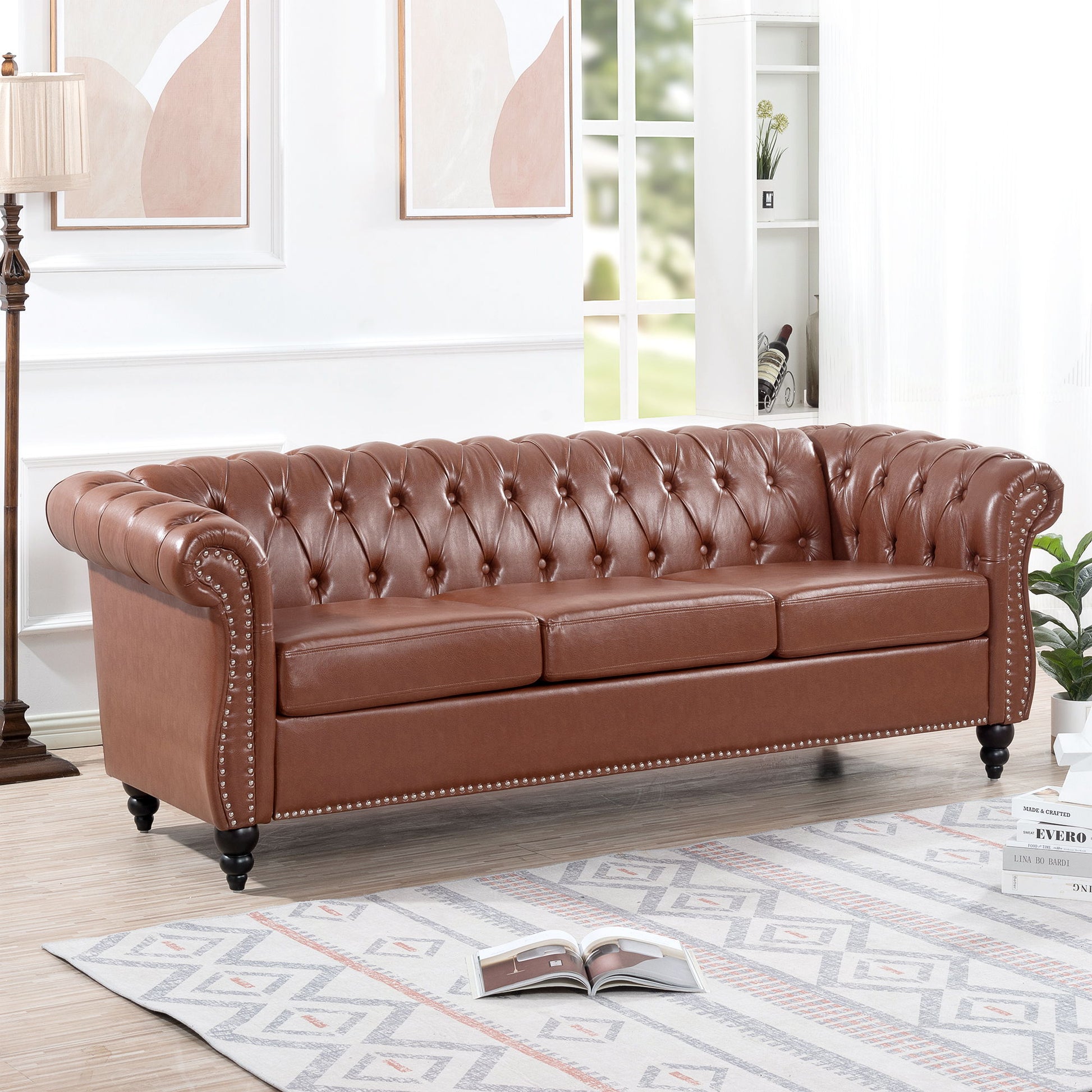 The Rolled Arm Chesterfield 3 Seater Sofa, featuring brown tufted leather and rolled arms, sits in a bright living room with modern decor, a patterned rug, framed art on the wall, and an open book on the floor.