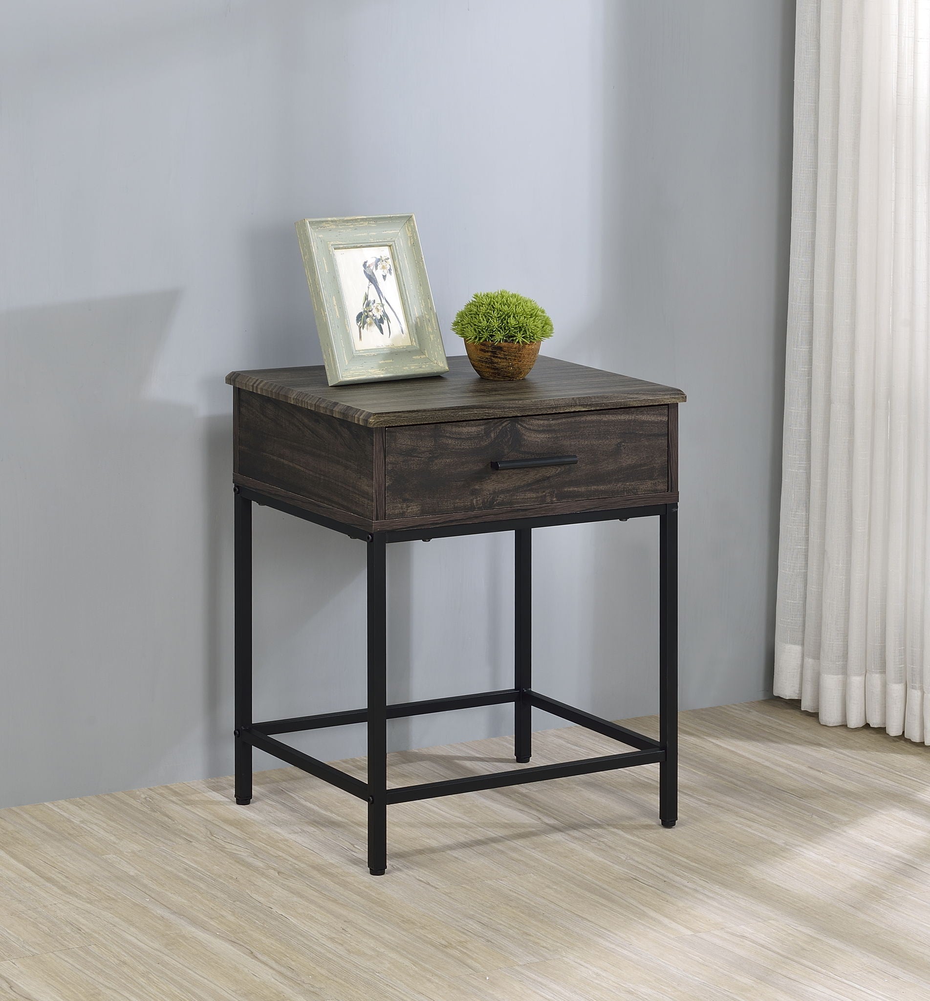 The Cliff - Modern Design Table with a drawer and black metal legs stands by a light gray wall. A framed photo and small green potted plant rest on top, complemented by light wood flooring and white curtains in the background.