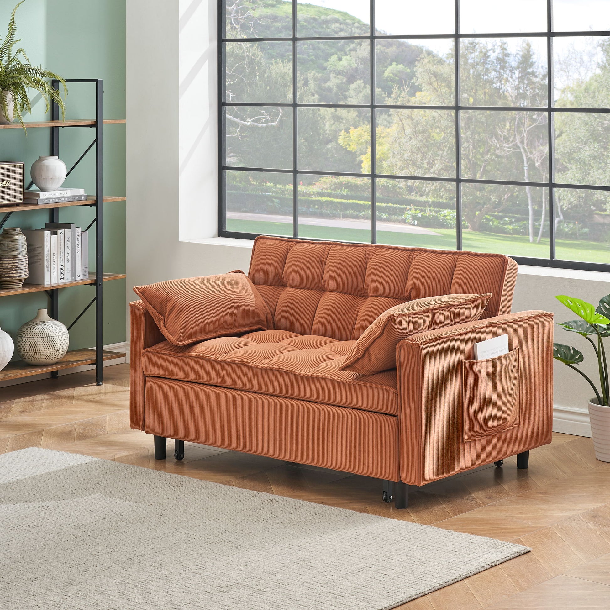 A modern living room features the Folding Sofa Bed with adjustable back—a rust-colored loveseat with tufted cushions, pillows, side pocket—set by a large window, bookshelf, green wall, wooden floor, and potted plant.