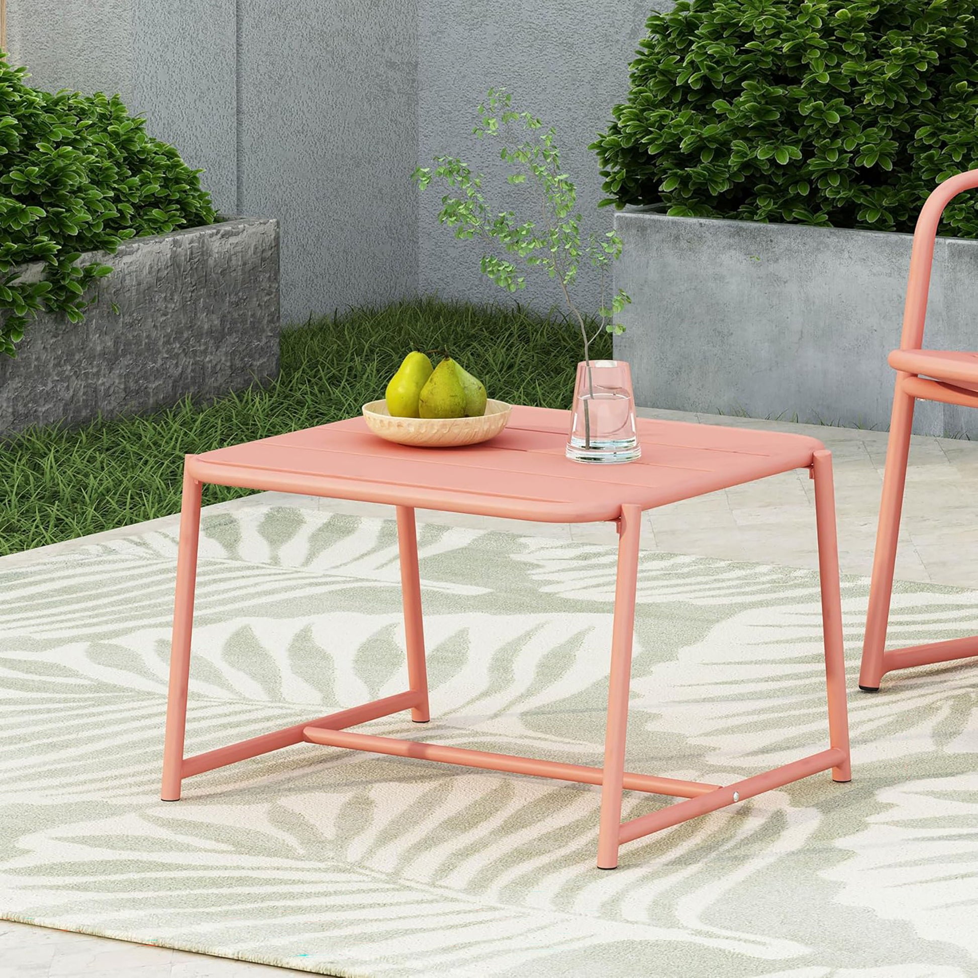 The Conrad - Side Table Outdoor in pink is styled with a bowl of pears and a glass of water topped with greenery, set on a patterned rug with green bushes and a gray planter in the background.