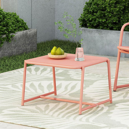 The Conrad - Side Table Outdoor in pink is styled with a bowl of pears and a glass of water topped with greenery, set on a patterned rug with green bushes and a gray planter in the background.
