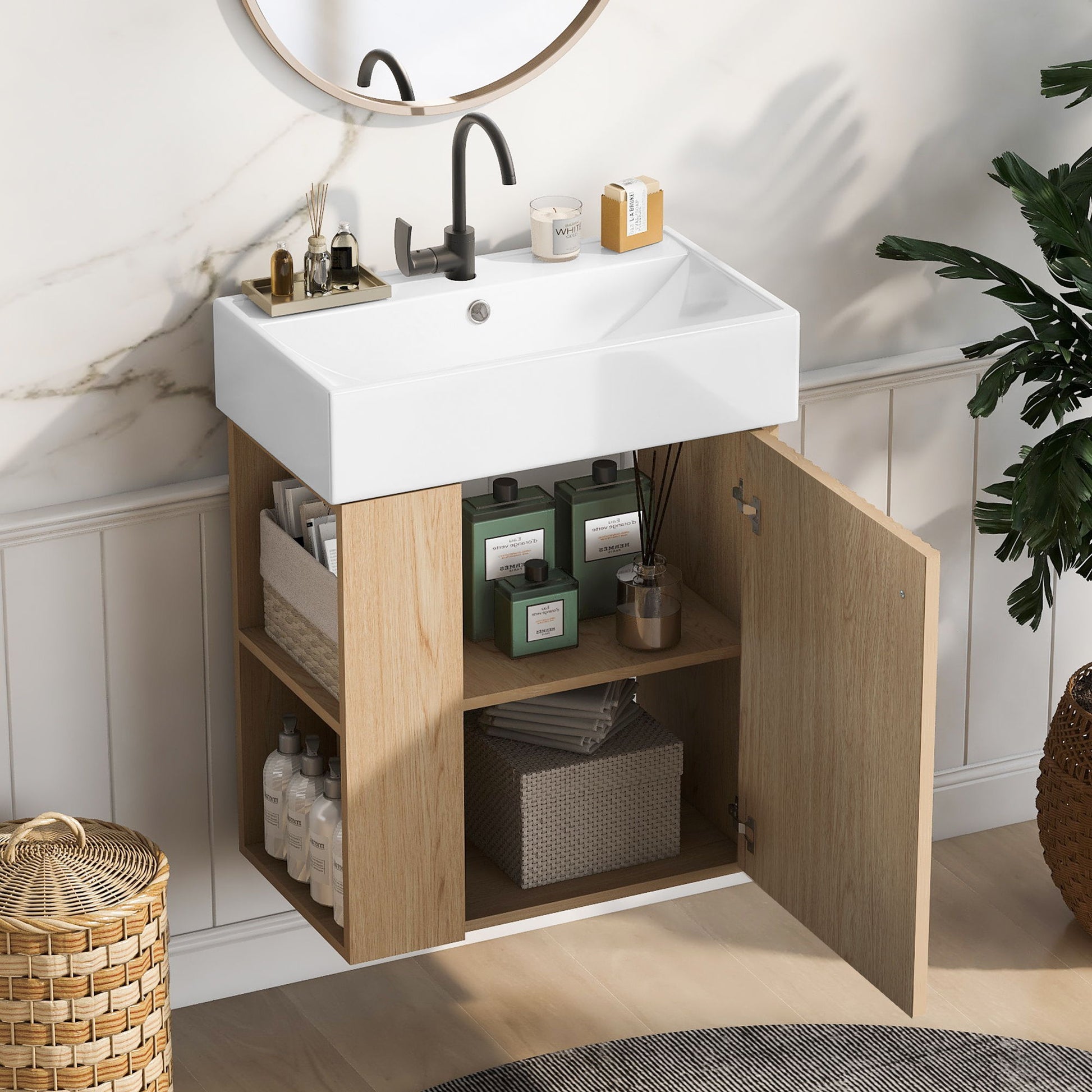 The Modern Floating Bathroom Vanity With Ceramic Basin features a white rectangular sink, black faucet, wooden cabinet with open shelves for bottles, storage for towels and boxes, plus a nearby round mirror and potted plant—ideal for small bathrooms.