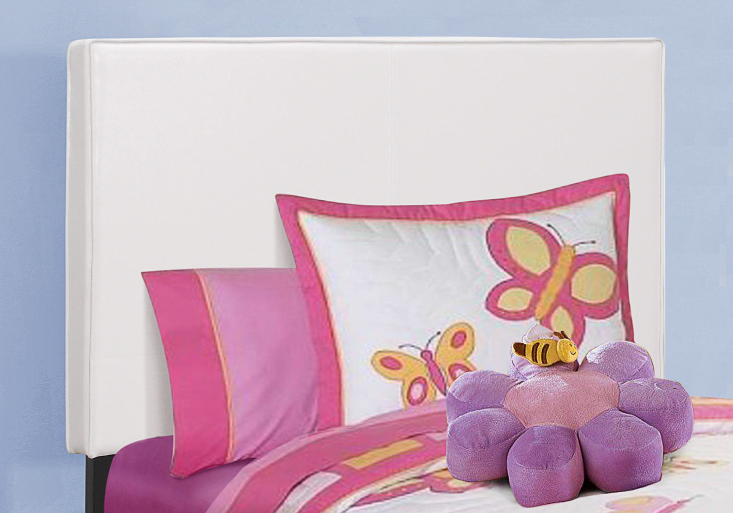 An upholstered transitional bed with pink and purple bedding, butterfly-print pillow, a plush purple flower, and a small bee toy sits against a light blue wall and white headboard.