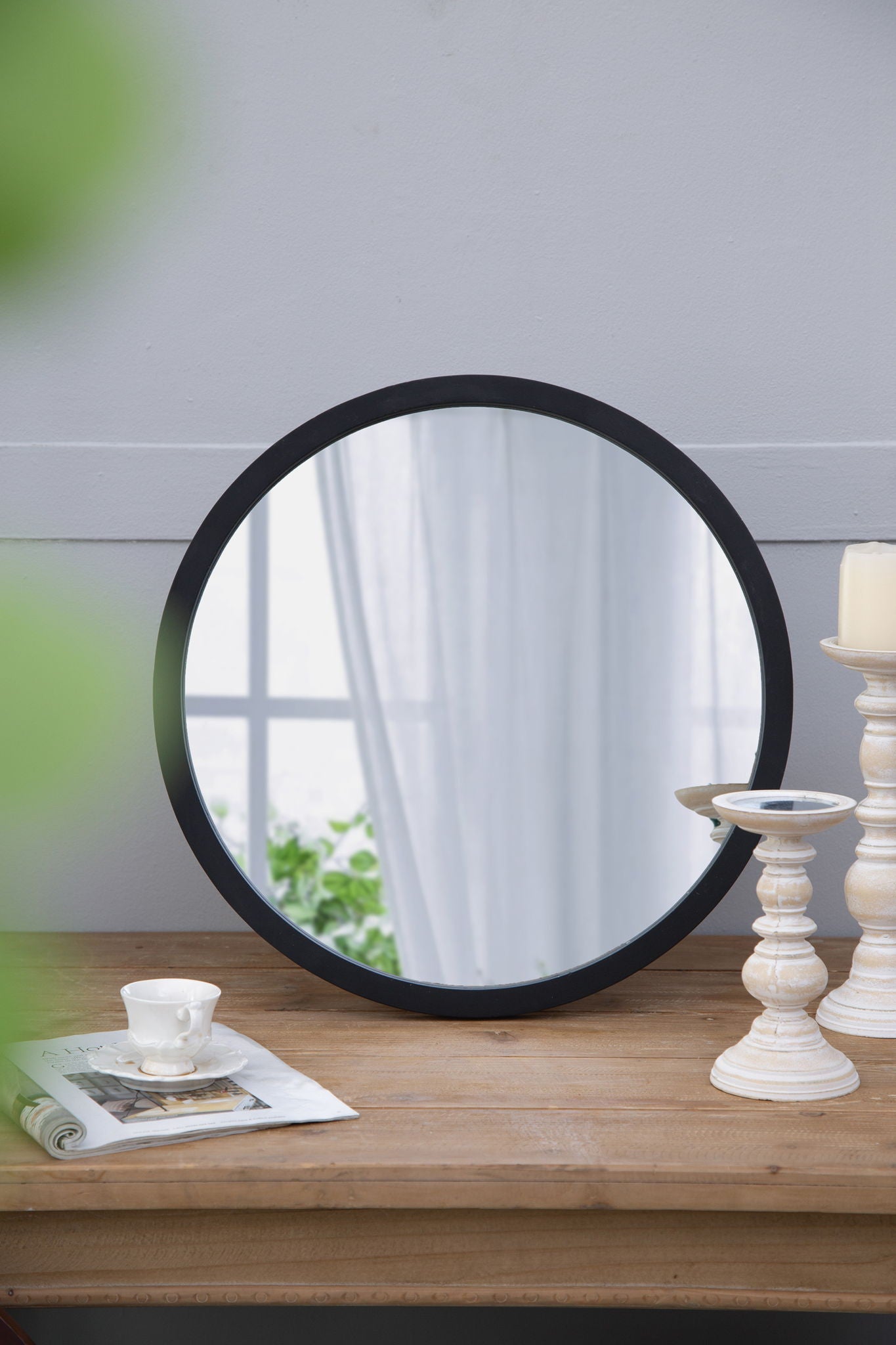 A Circle Wall Mirror With Wooden Frame rests on a wooden table beside white candlesticks, a teacup with saucer, and an open magazine; its reflection shows a window with sheer curtains and greenery.