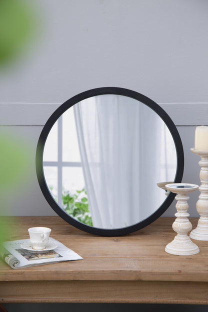 A Circle Wall Mirror With Wooden Frame rests on a wooden table beside white candlesticks, a teacup with saucer, and an open magazine; its reflection shows a window with sheer curtains and greenery.