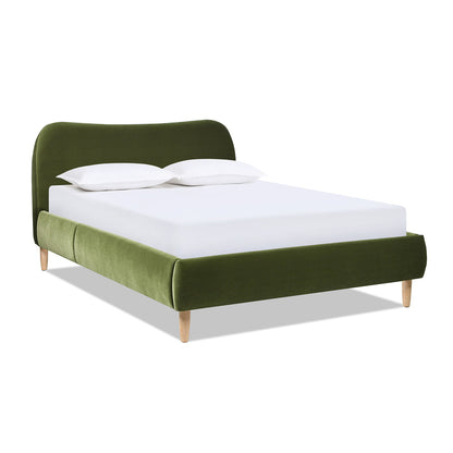 The Roman - Curved Headboard Upholstered Platform Bed features green velvet upholstery, light wooden legs, a curved headboard, and a low-profile footboard. Shown with a white mattress and two white pillows.