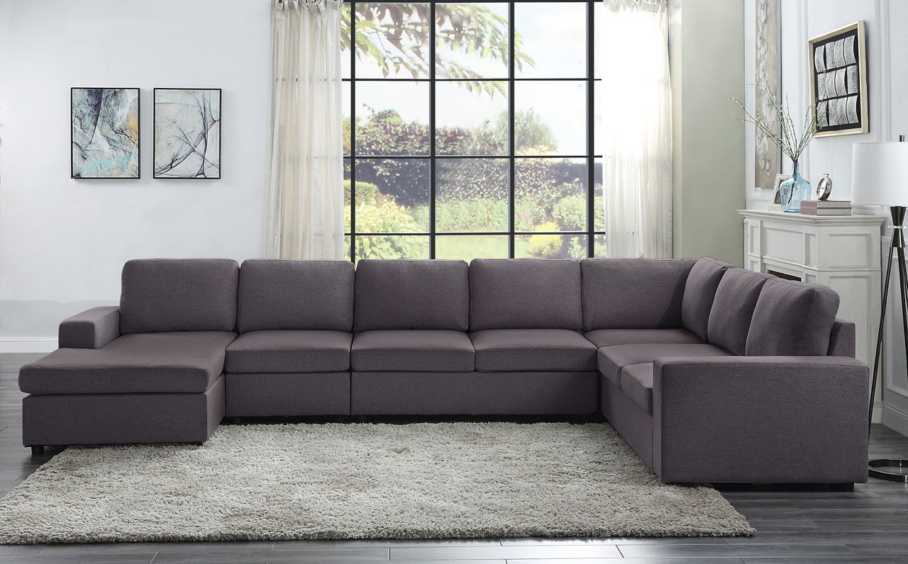 A modern living room with the Tifton - Linen 7 Seat Reversible Modular Sectional Sofa Chaise in gray on a light rug, featuring a wall of windows, sheer curtains, abstract art, and a floor lamp beside a white console table.