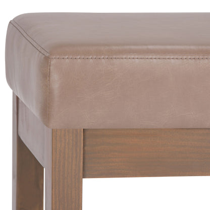 A close-up of the Milltown Upholstered Ottoman Bench, featuring a padded light brown leather seat with visible edge stitching and medium brown wooden legs.