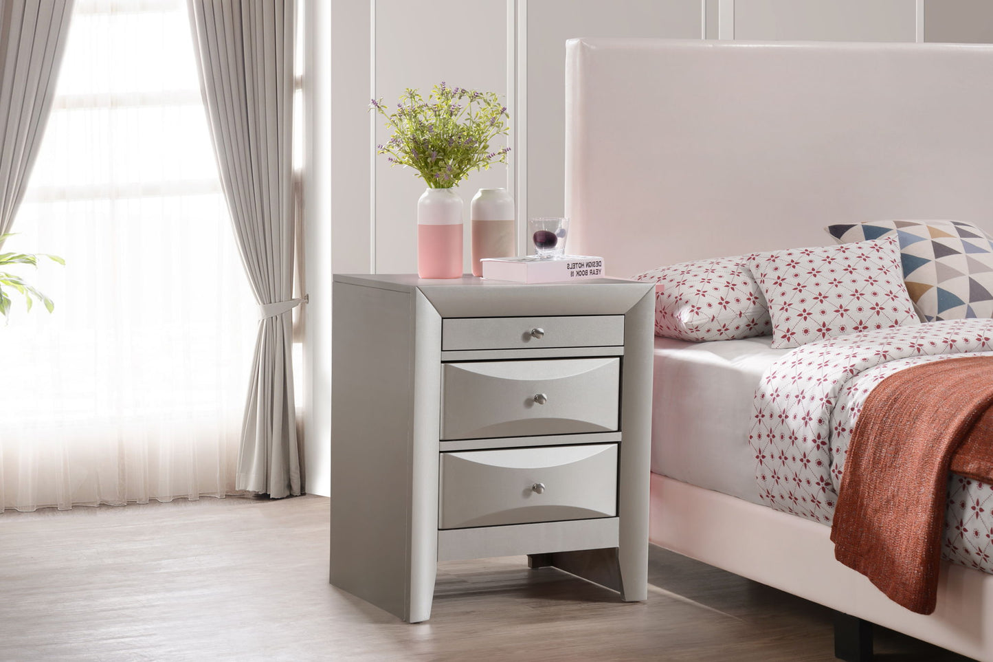 A modern bedroom features the Marilla Nightstand in white with three drawers, adorned with a vase of green flowers, two decorative jars, a book, and a wine glass beside a bed with patterned bedding and an orange blanket.