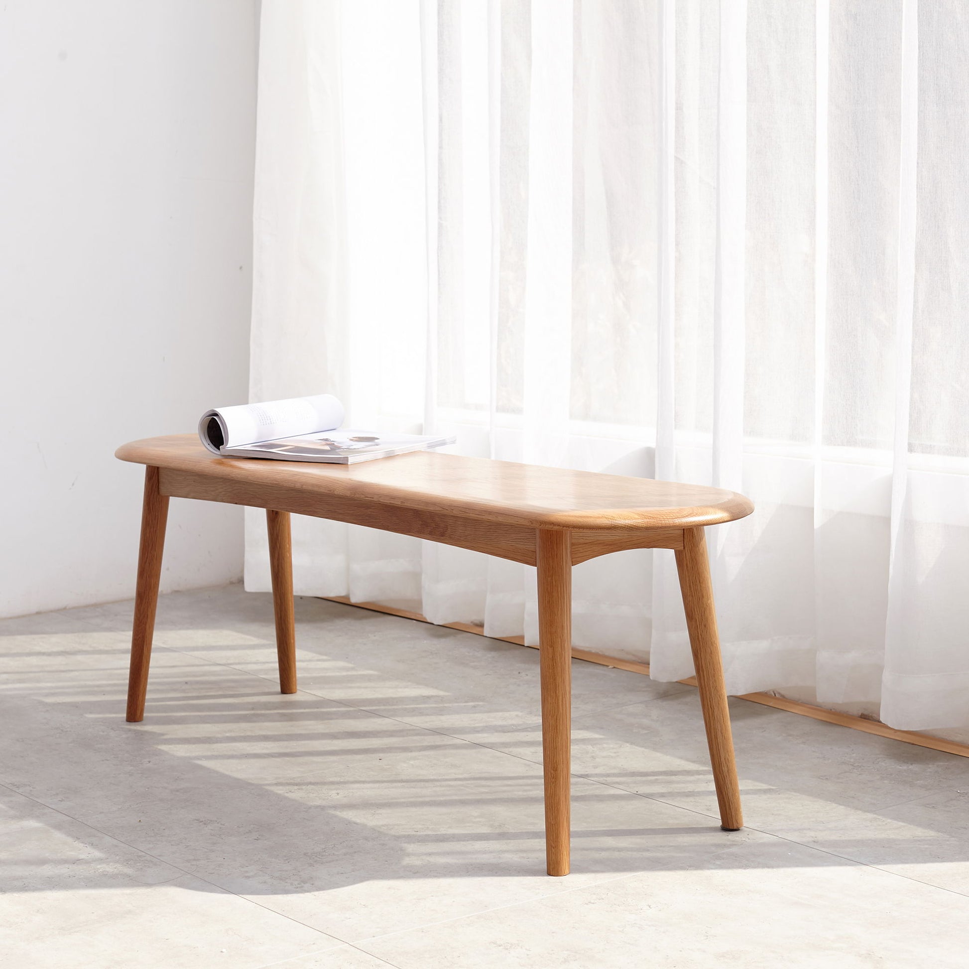 The Dining Bench, Table Bench For Living Room with four legs sits on a sunlit floor by white curtains. An open magazine lies on the bench as soft natural light fills the minimalist space.