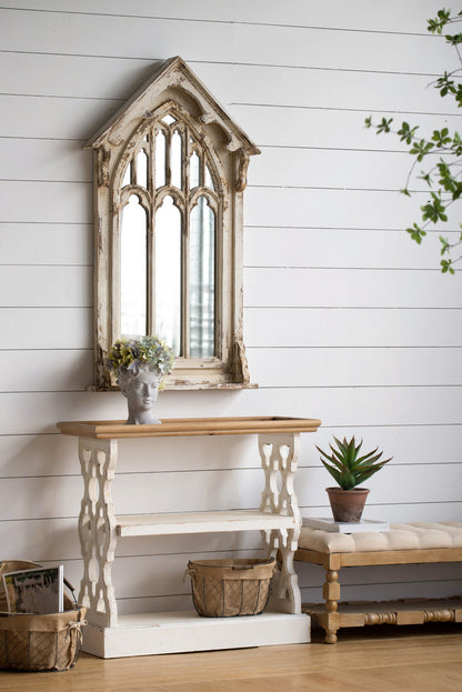 A rustic entryway features a Gothic arched mirror, a Distressed Wood Shelf Tray French Country Console Table in brown/antique white with a flower-holding bust, baskets, a potted succulent, a woven bench, and white shiplap walls.