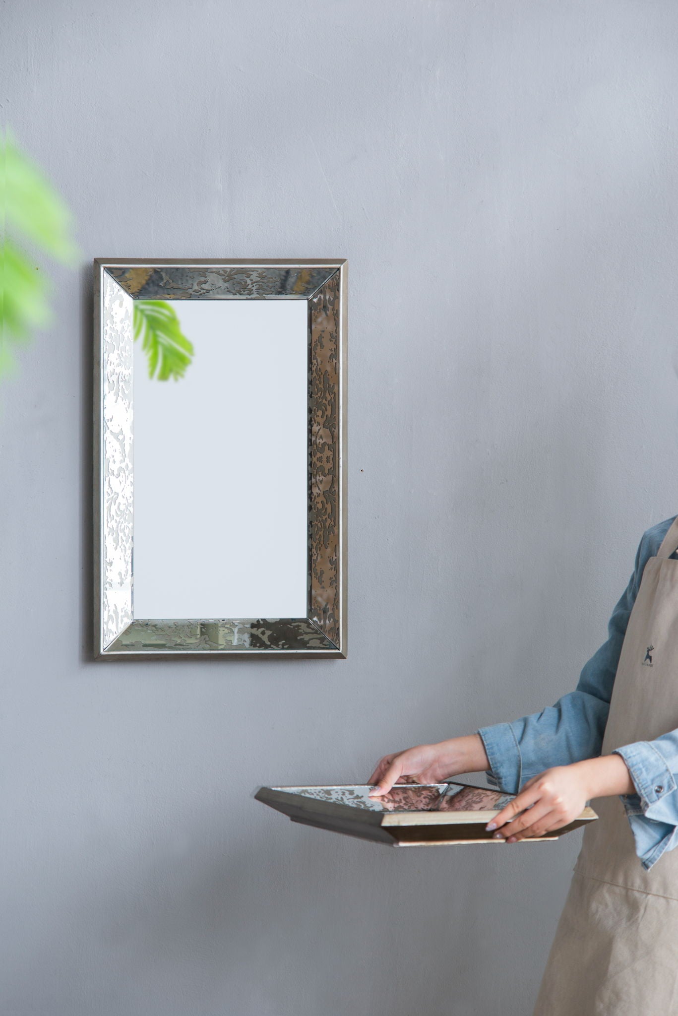 A person in a blue shirt and beige apron holds a food tray near the Antique Rectangle Mirror With Floral Accents, a silver-framed hanging wall mirror, with part of a green plant visible on the left against a gray wall.