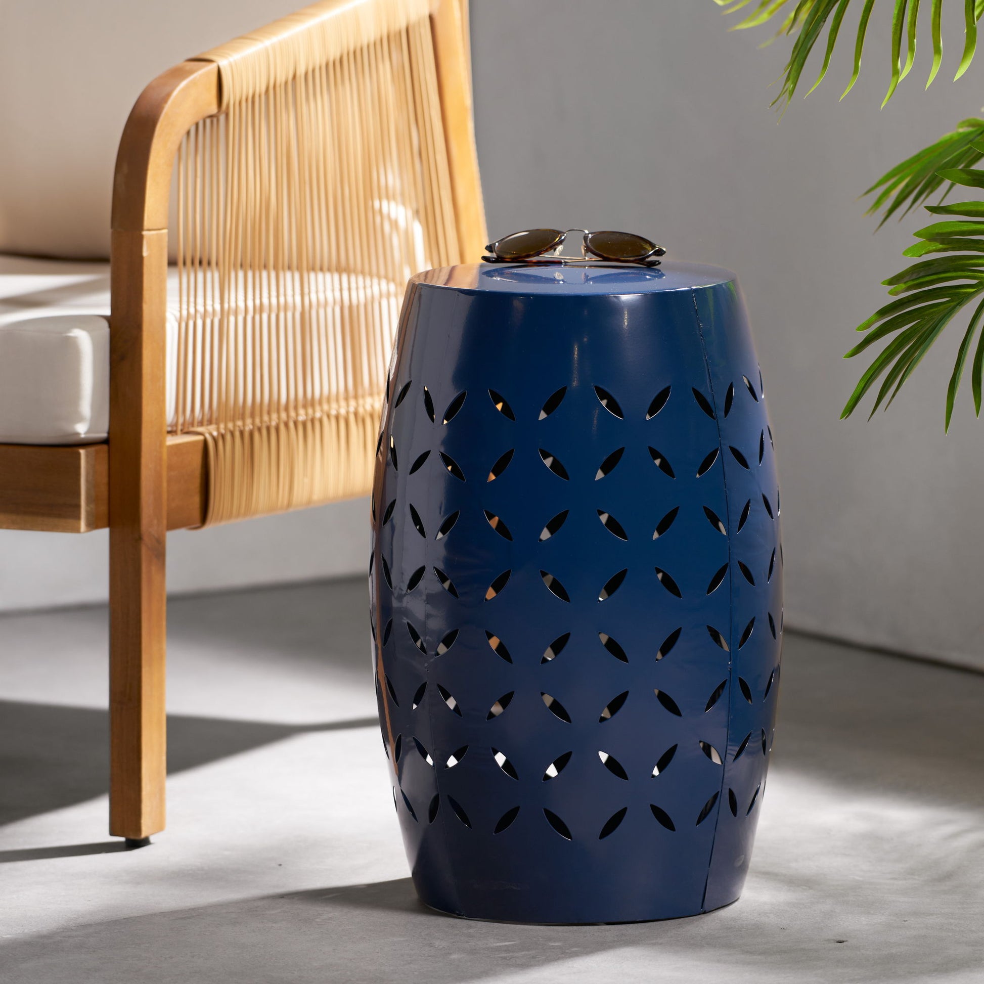 A 12" Outdoor Lace Cut Side Table in blue metal with a leaf pattern sits by a rattan chair with a white cushion. Sunglasses are on the table, and a green plant is visible to the right.