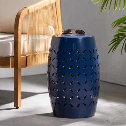 A 12" Outdoor Lace Cut Side Table in blue metal with a leaf pattern sits by a rattan chair with a white cushion. Sunglasses are on the table, and a green plant is visible to the right.