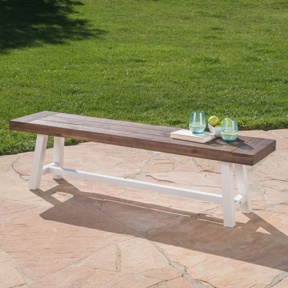 The Tranquilizer Outdoor Acacia Wood Bench With Slat Panel Design, featuring white legs, is placed on a stone patio with two glasses of water, a small white plate, and three green apples. A lush lawn and trees can be seen in the background.