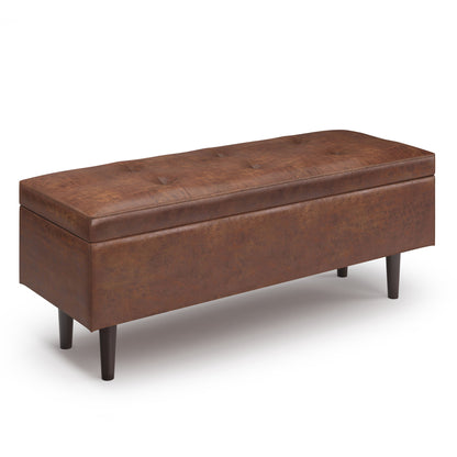 The Shay Large Square Coffee Table Storage Ottoman features brown faux leather upholstery with a tufted lid and four dark wood legs, shown on a white background. Mid-century style design adds functional elegance to any room.