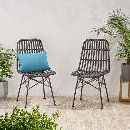 Two Sawtelle black wicker chairs with metal legs sit side by side on a light stone floor. One chair features a blue cushion, and green potted plants are arranged around them against a white wall.