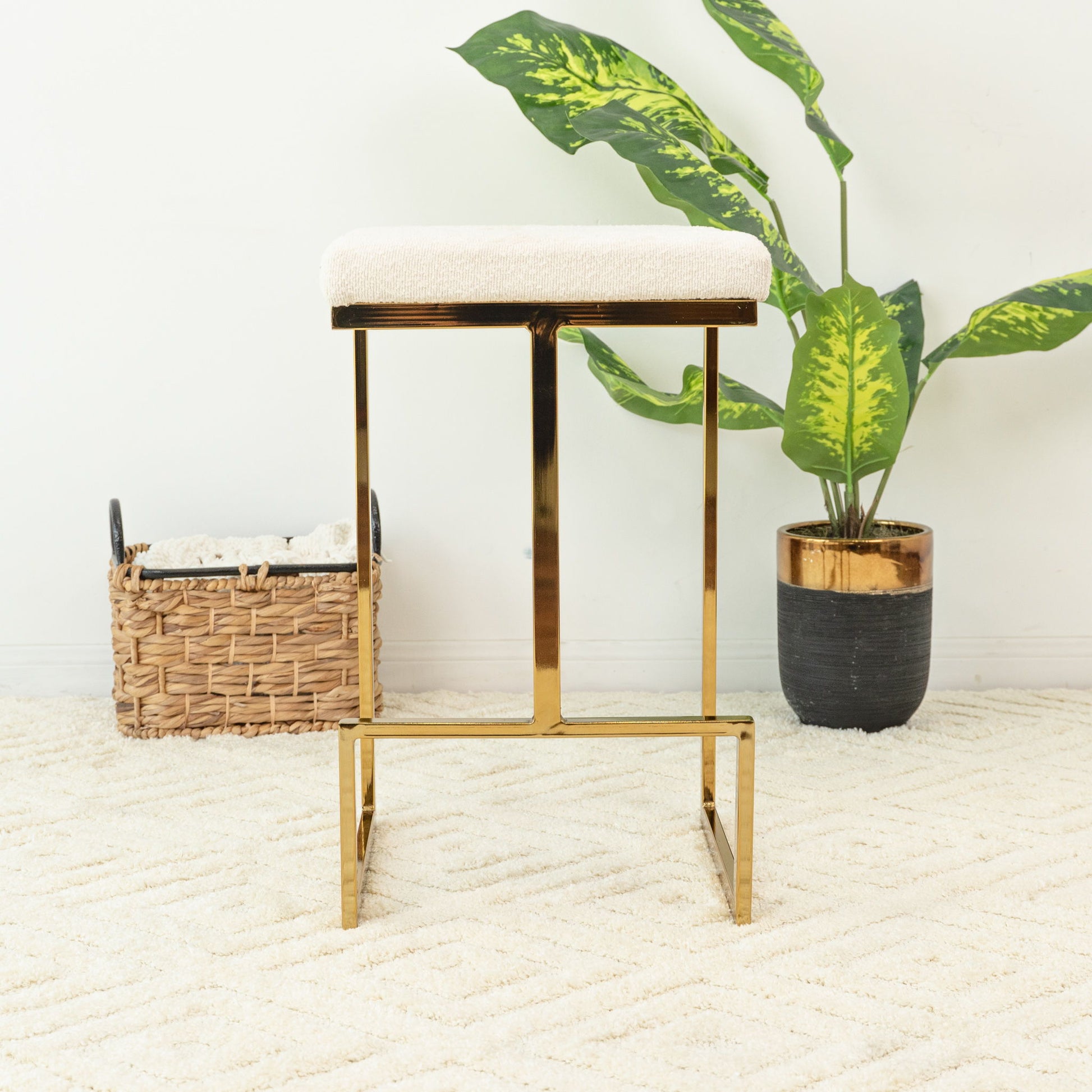 The Joel - Mid Century Modern Luxury Upholstered Stool, featuring a gold metal frame and white cushion, sits on a plush cream rug with a green potted plant and wicker basket in the background against a plain white wall.