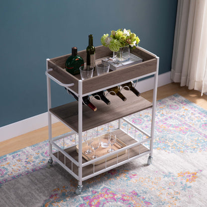 The Rolling Kitchen Cart With Storage And Four Wine Bottle Rack displays wine bottles, liquor, glasses, and a vase with green flowers. It's set on a colorful rug beside a blue wall and a window with light curtains.