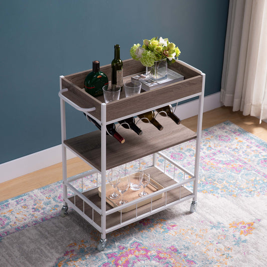 The Rolling Kitchen Cart With Storage And Four Wine Bottle Rack displays wine bottles, liquor, glasses, and a vase with green flowers. It's set on a colorful rug beside a blue wall and a window with light curtains.