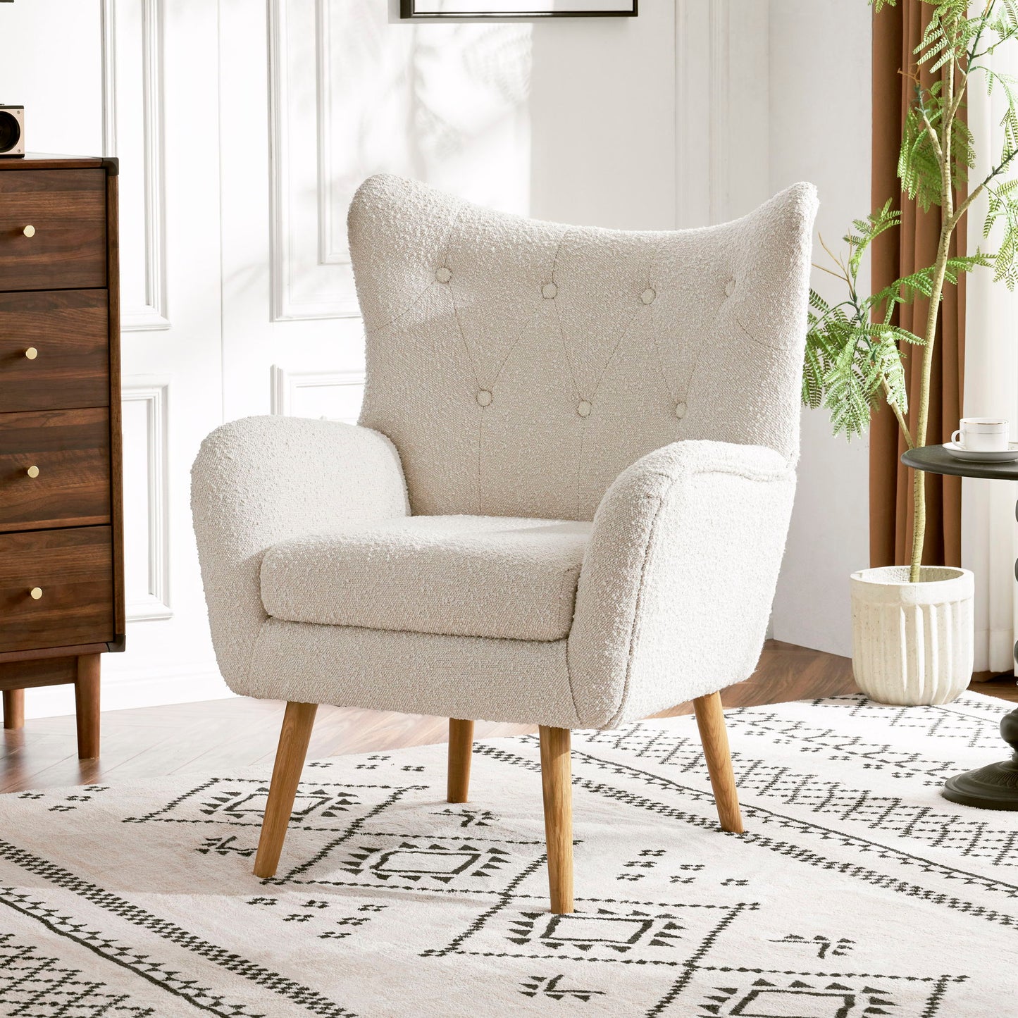 The Mid-Century Modern Arm Chair with wingback design and wood legs, upholstered in white, sits on a patterned rug in a bright room featuring white walls, a wooden dresser, round table, and potted plant.