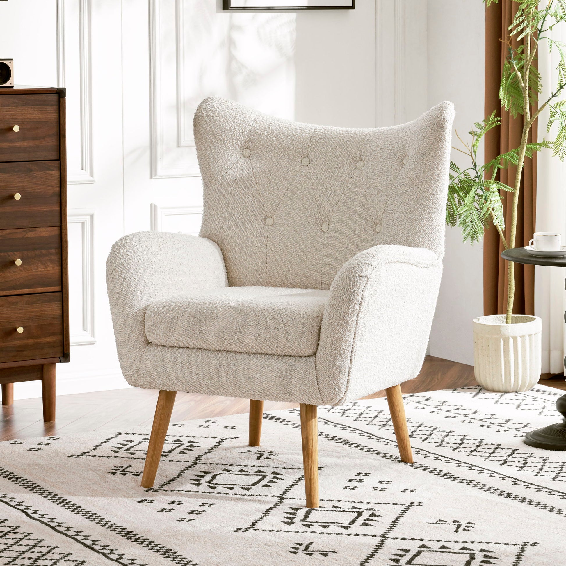 The Mid-Century Modern Arm Chair with wingback design and wood legs, upholstered in white, sits on a patterned rug in a bright room featuring white walls, a wooden dresser, round table, and potted plant.