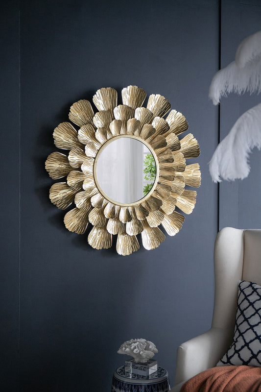 The Round Metal Mirror With Trumpet Vine Motif - Gold hangs on a dark wall above a small blue table, with a cream armchair and white feathery decor piece partially visible in the foreground.