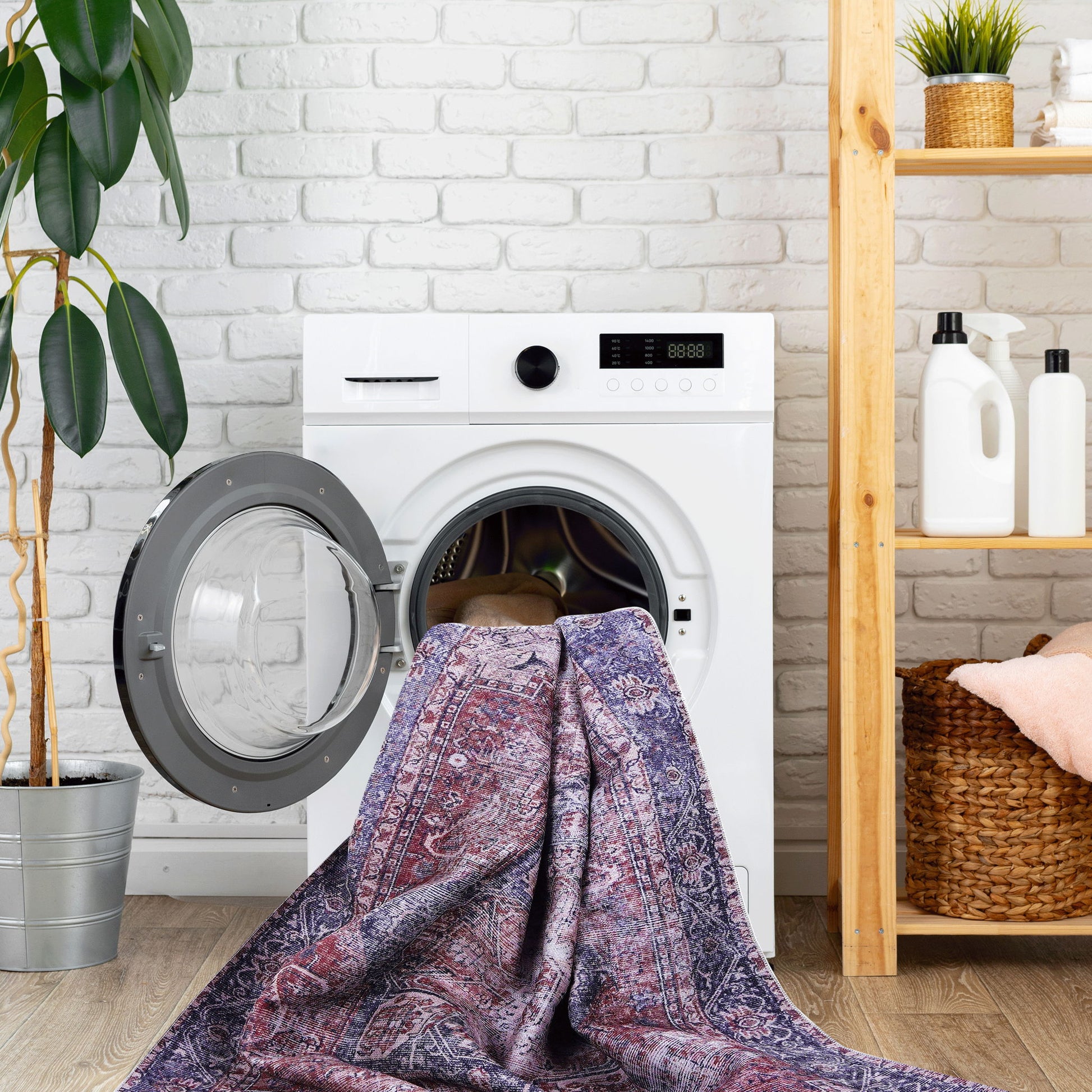 A Maval - Unique Washable Area Rug is partly inside an open washing machine, set by a white brick wall with a plant and wooden shelves holding laundry supplies and towels.