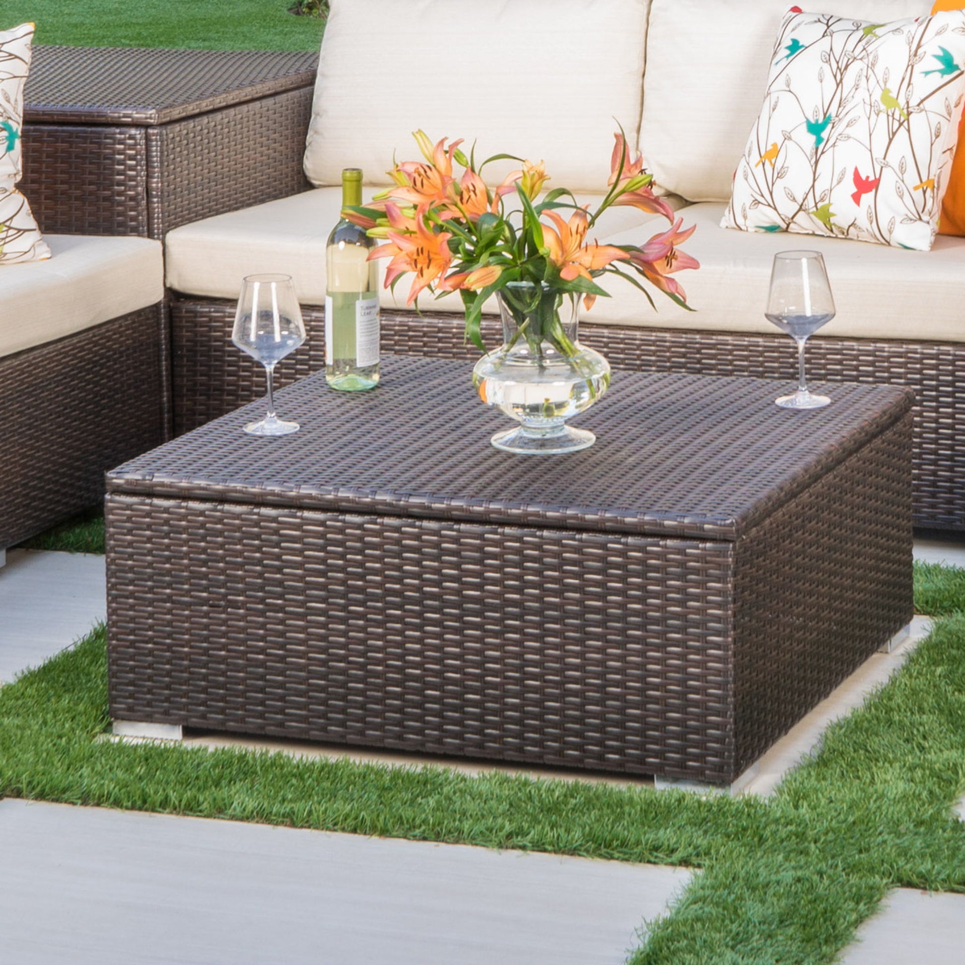 The Santa Rosa - Storage Table, adorned with a glass vase of orange lilies, a wine bottle, and two glasses, stands on a patio surrounded by cushioned seats with patterned pillows and green grass.