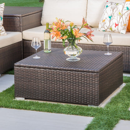 The Santa Rosa - Storage Table, adorned with a glass vase of orange lilies, a wine bottle, and two glasses, stands on a patio surrounded by cushioned seats with patterned pillows and green grass.
