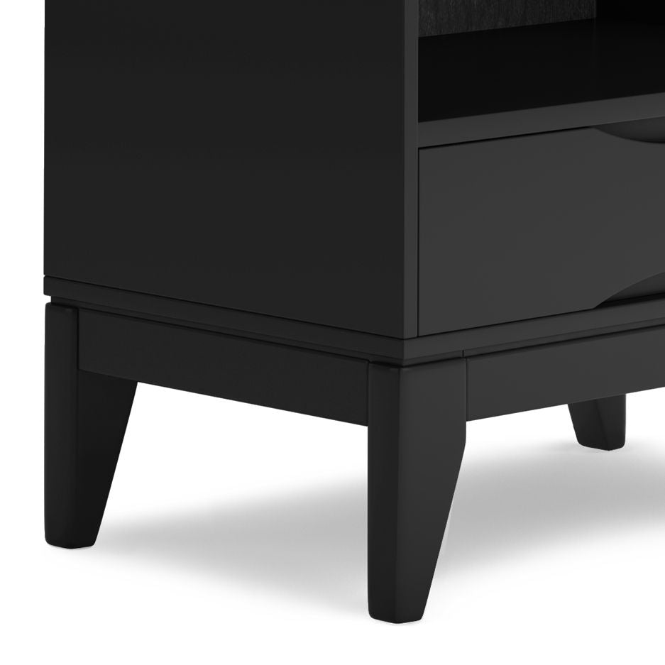 A close-up view of the bottom corner of the black Harper - Handcrafted Bookcase With Storage, featuring tapered legs, an open shelf, and a storage drawer.