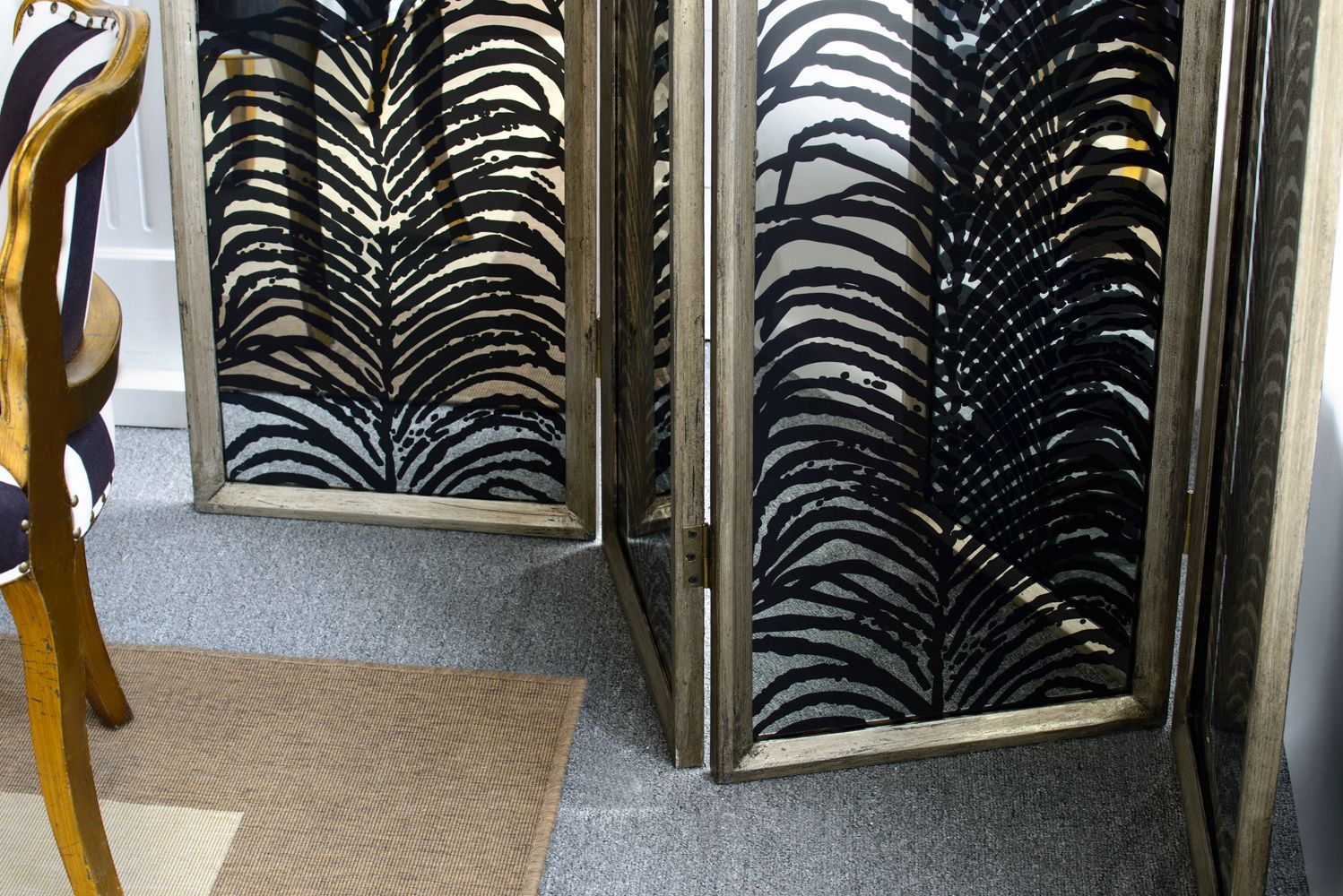 The Herero 4 Panel Decorative Screen - Black features palm leaf patterns on glass panels and stands on a gray carpet beside a wooden chair and brown rug.