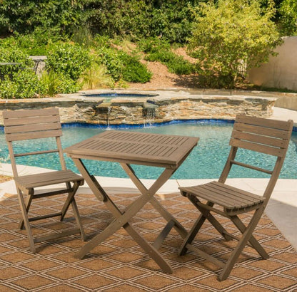 The Positano Foldable Dining Set, featuring two chairs and a square table, is arranged on a patterned outdoor rug beside a backyard pool with lush greenery and a stone waterfall in the background.