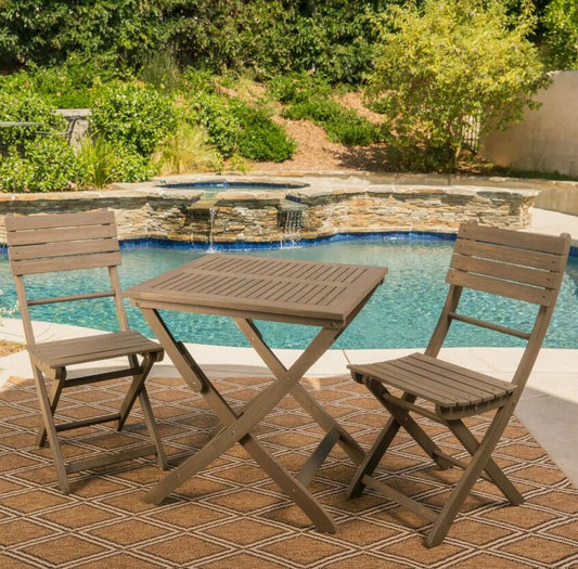 The Positano Foldable Dining Set, featuring two chairs and a square table, is arranged on a patterned outdoor rug beside a backyard pool with lush greenery and a stone waterfall in the background.