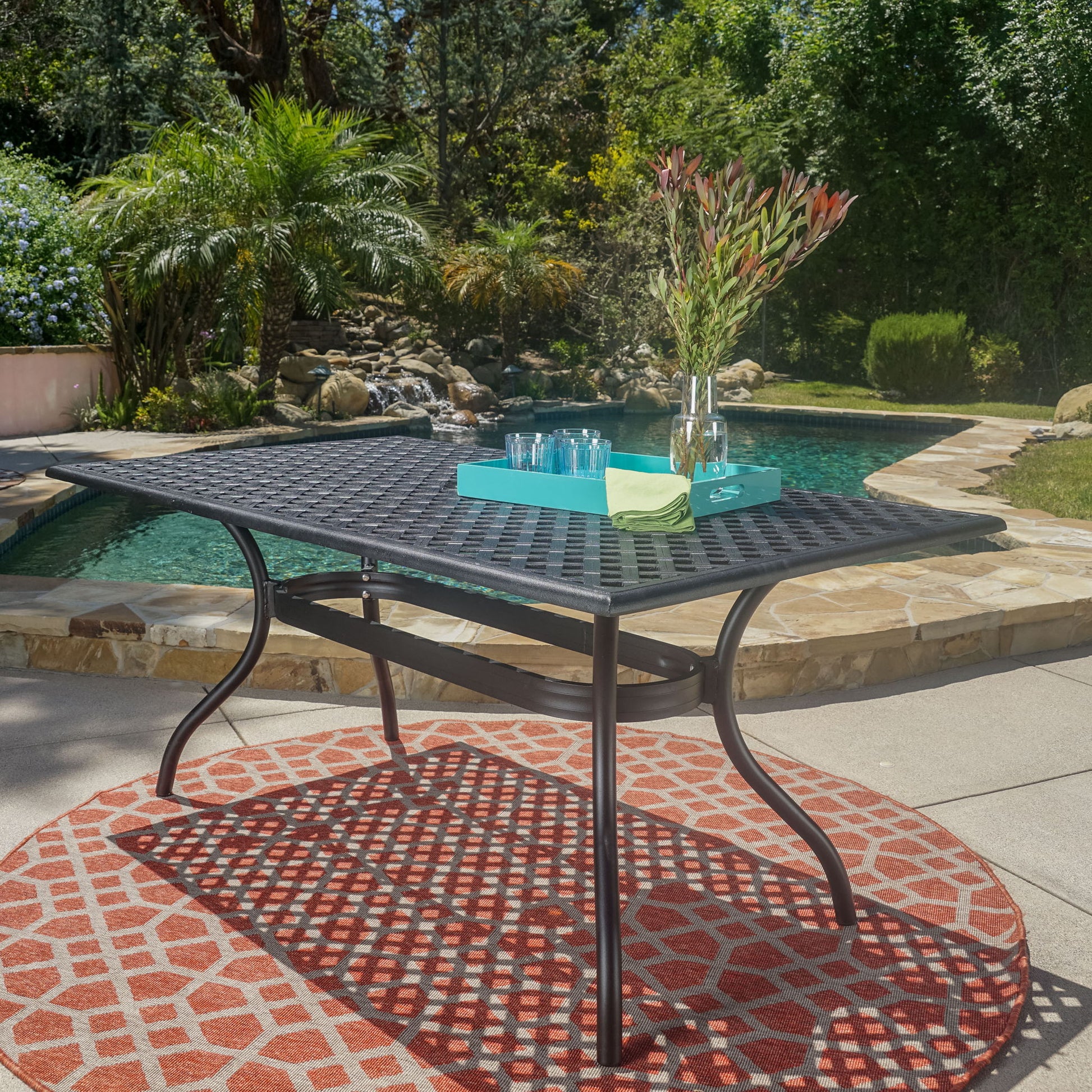 The Outdoor Aluminum Dining Table With Lattice Design And Center Umbrella Hole - Black sits on a red patterned rug beside a pool, topped with a blue tray, glassware, green napkin, and flower vase. The pool is surrounded by lush greenery.