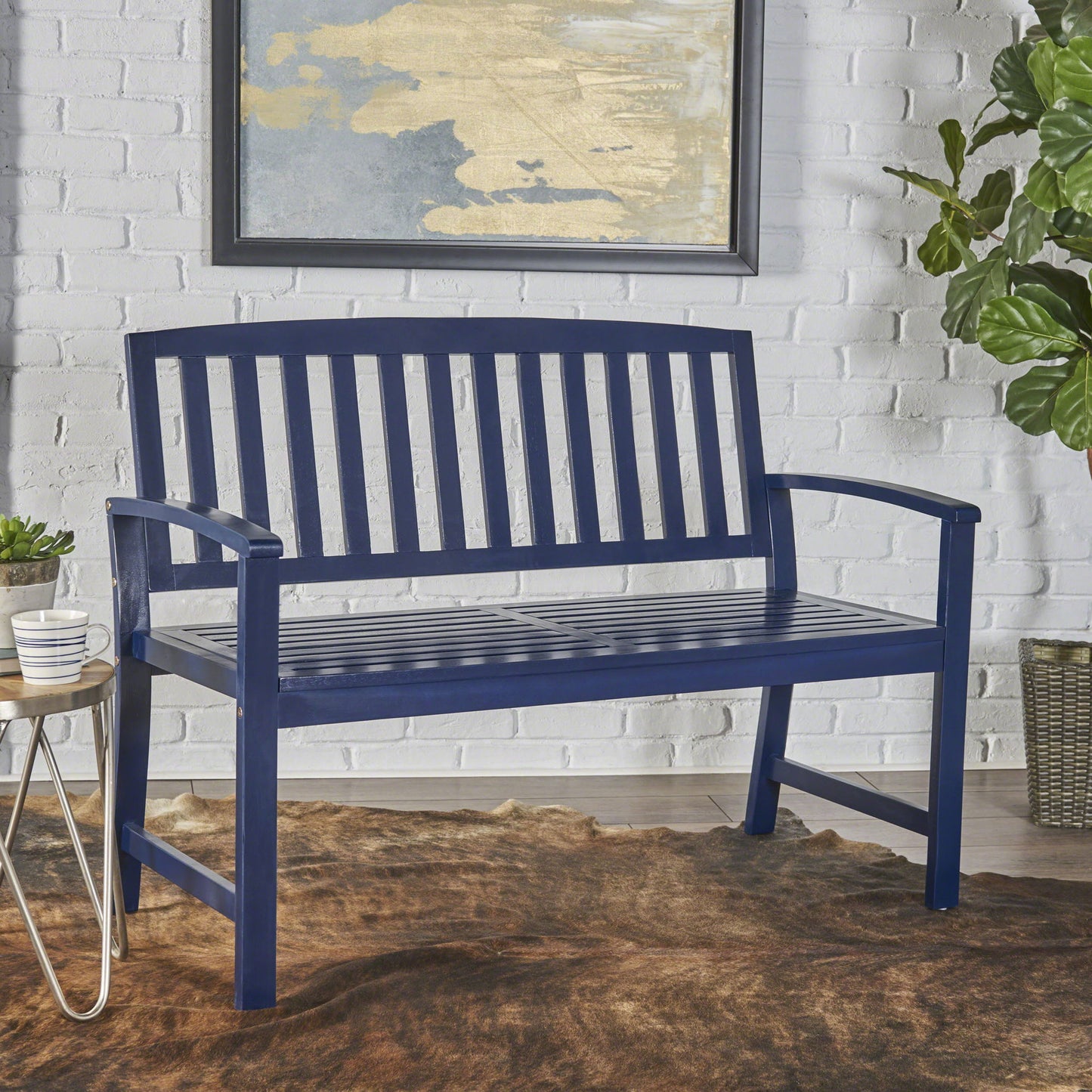 The Farmhouse Style Loja Bench Indoor / Outdoor, finished in blue wood with vertical slats, stands against a white brick wall. A modern painting hangs above, with plants and a side table nearby on a brown rug.