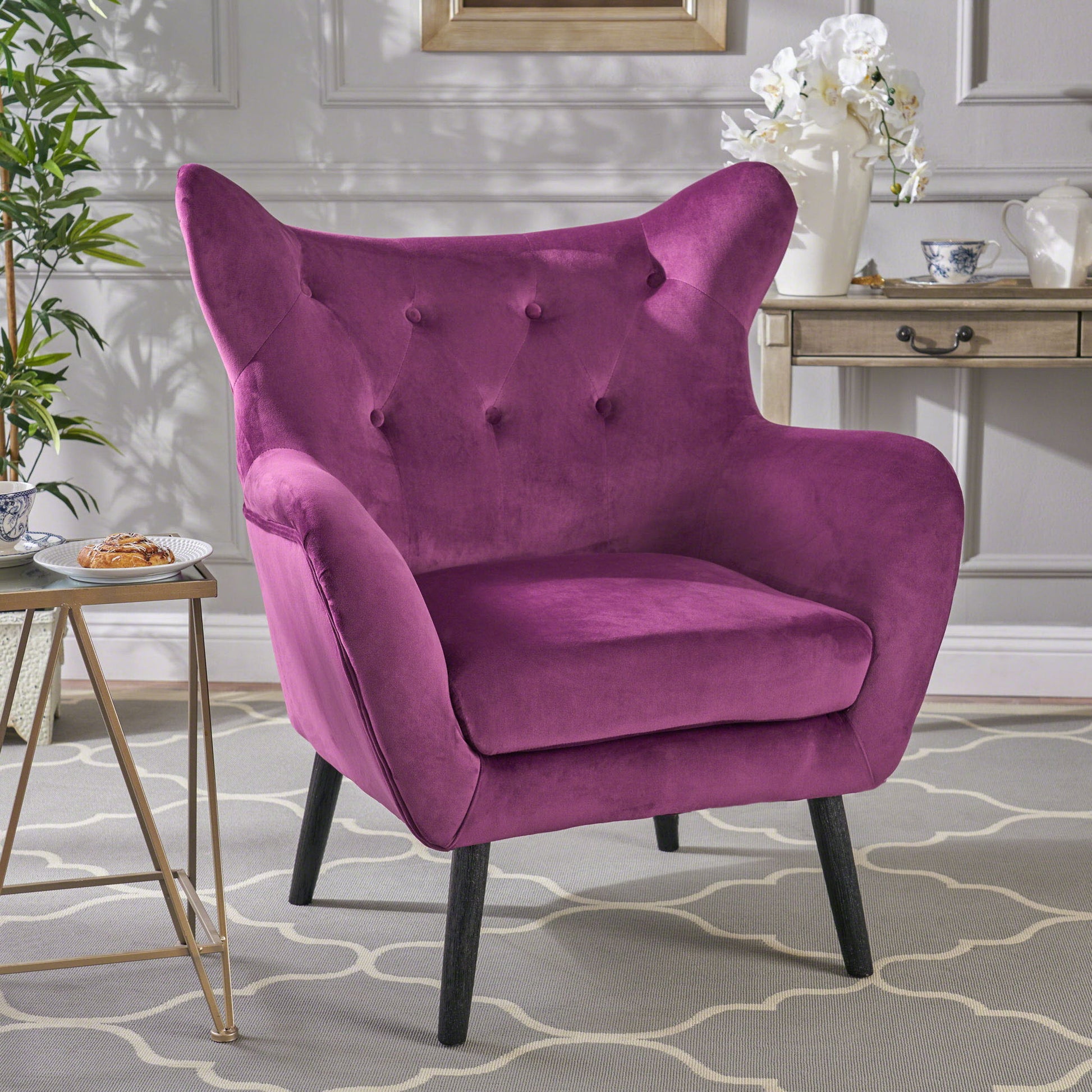 The Alyssa Mid Century Upholstered Arm Chair with wingback design, featuring plush magenta tufted upholstery and black legs, sits on a patterned rug next to a side table in a stylish room with light gray walls.