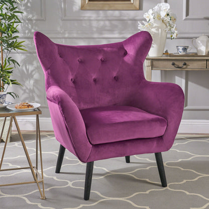 The Alyssa Mid Century Upholstered Arm Chair with wingback design, featuring plush magenta tufted upholstery and black legs, sits on a patterned rug next to a side table in a stylish room with light gray walls.