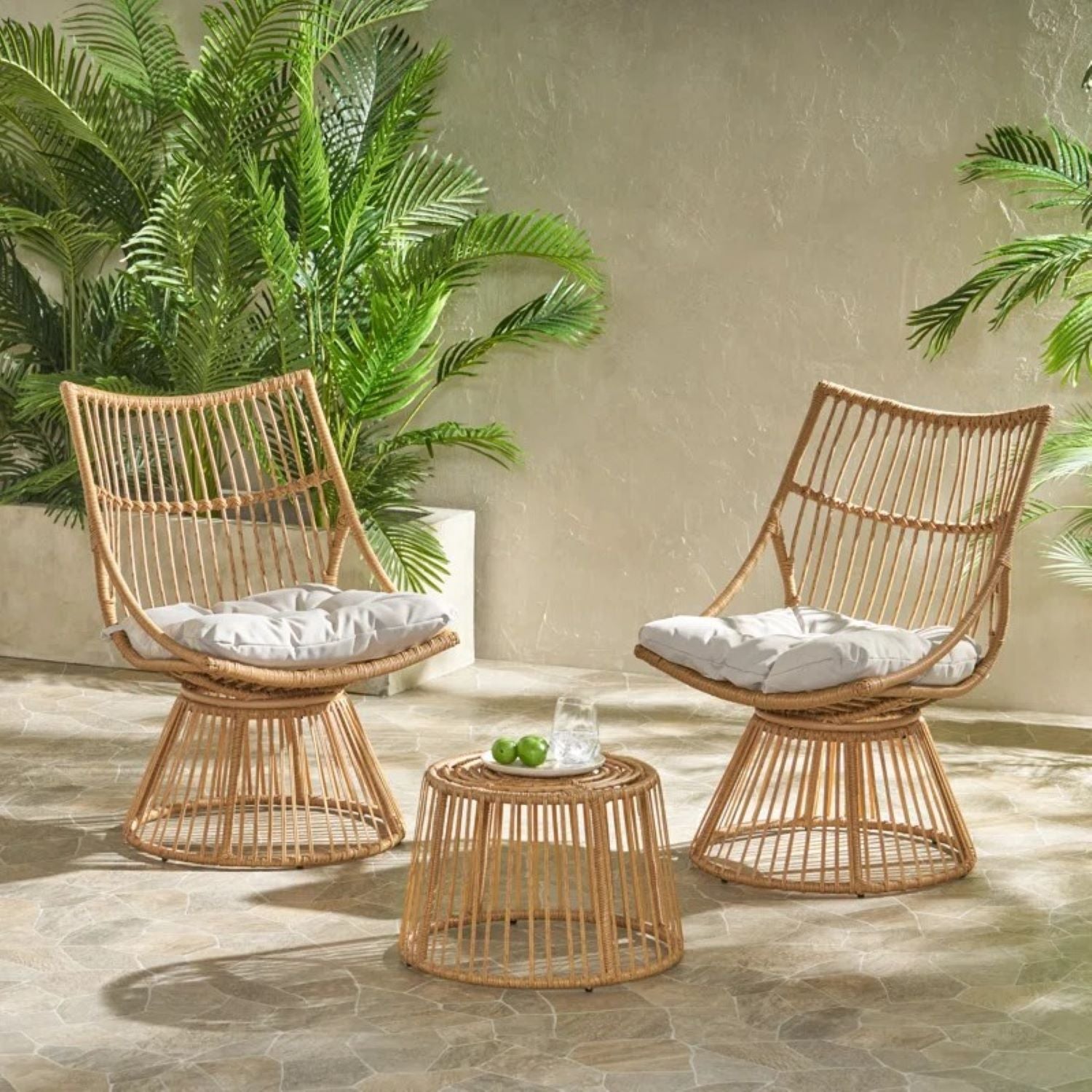 The Elaine - Chair & Side Table set, featuring two wicker chairs with white cushions and a round wicker table, sits on a stone patio surrounded by green potted plants against a light wall, with a glass of water and limes on the table.