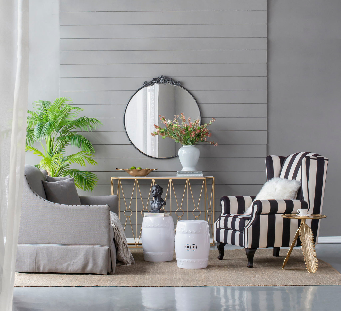 A modern living room with a gray sofa, striped armchair, gold console table, white stools, neutral décor, and a Classic Design Mirror with Baroque inspired frame leaning against the wall on a woven rug.