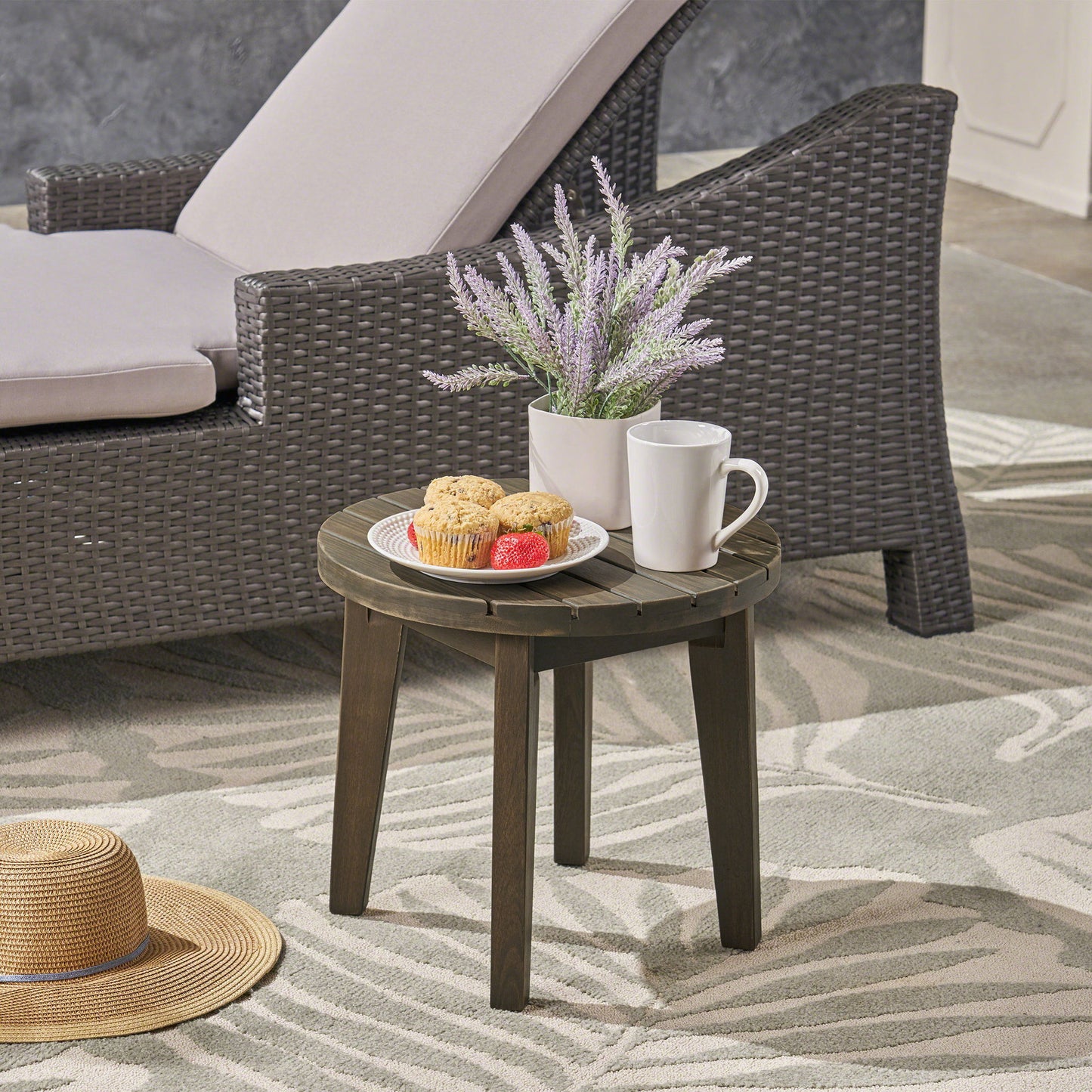 The Outdoor Acacia Wood Side Table With Weather Resistance holds muffins, strawberries, a white mug, and lavender in a white pot beside a wicker lounge chair; a straw hat rests on the patterned outdoor rug nearby.