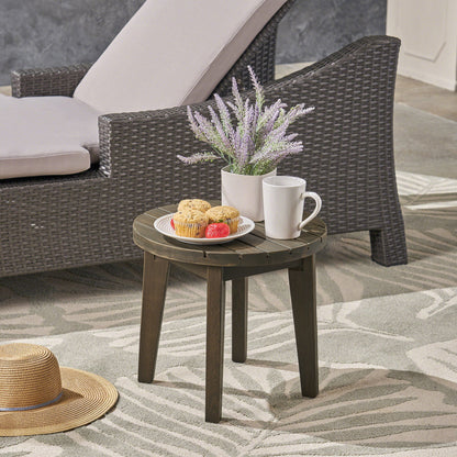 The Outdoor Acacia Wood Side Table With Weather Resistance holds muffins, strawberries, a white mug, and lavender in a white pot beside a wicker lounge chair; a straw hat rests on the patterned outdoor rug nearby.
