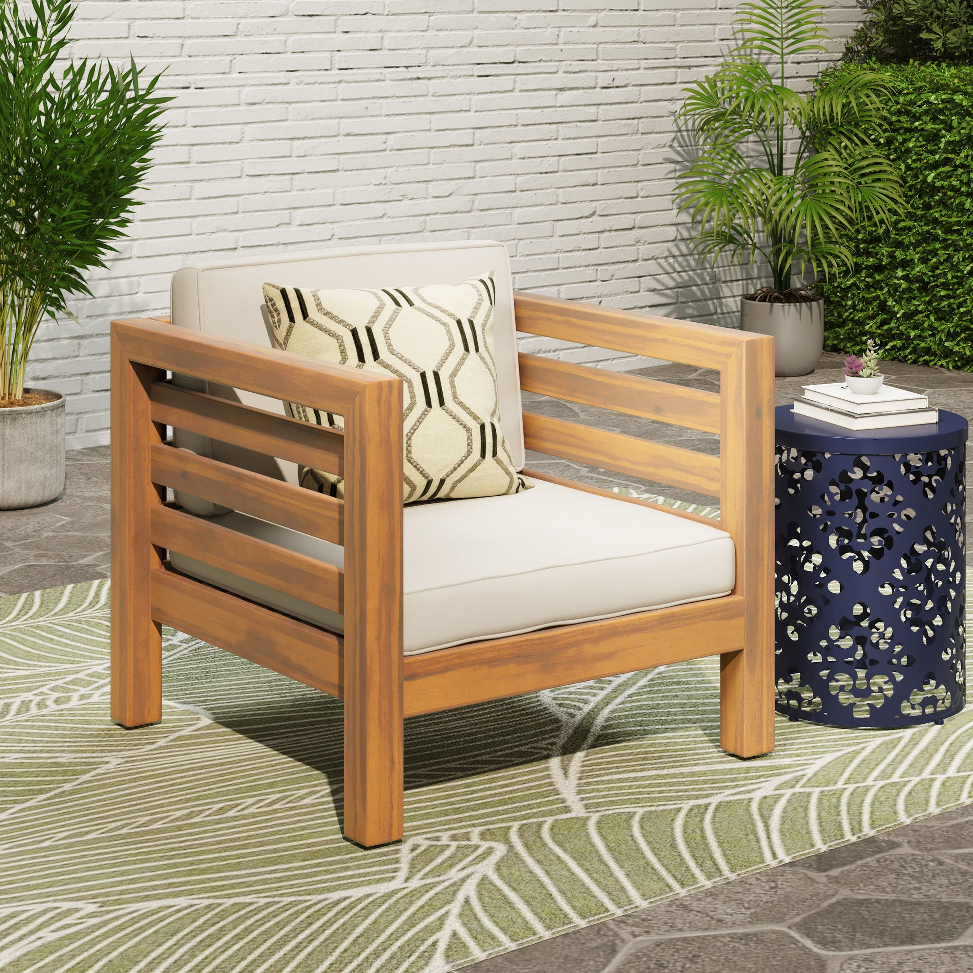 An Outdoor Acacia Wood Patio Chair With Fabric Cushion sits on a green leaf-patterned rug next to a blue side table, accented by potted plants and a geometric pillow against a white brick wall.