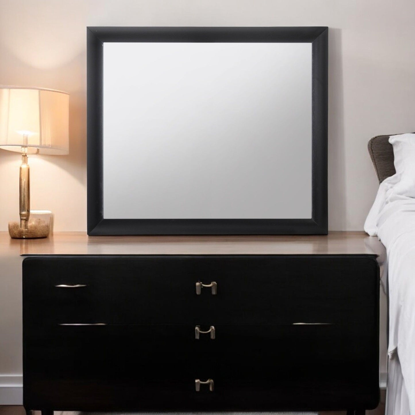 The Ireland - Contemporary Design Mirror with a black frame sits atop a dark wooden dresser next to a bed, alongside a lit table lamp with a beige shade on the left, set against a light-colored wall.