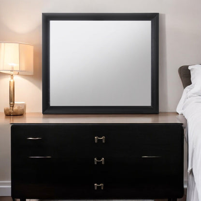 The Ireland - Contemporary Design Mirror with a black frame sits atop a dark wooden dresser next to a bed, alongside a lit table lamp with a beige shade on the left, set against a light-colored wall.