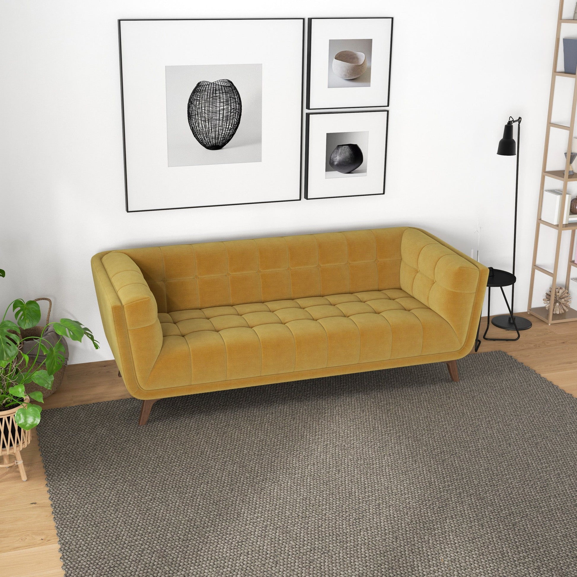 The Addison Mid-Century Modern Tufted Sofa in mustard yellow sits on a gray rug in a modern living room with wooden floors, a black floor lamp, minimalist framed art, a potted plant, and a wooden shelf.