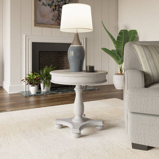 The Mariano Round End Table in gray sits beside a light gray sofa in a living room decorated with plants, a fireplace, framed art on the wall, and topped with a blue and white lamp.